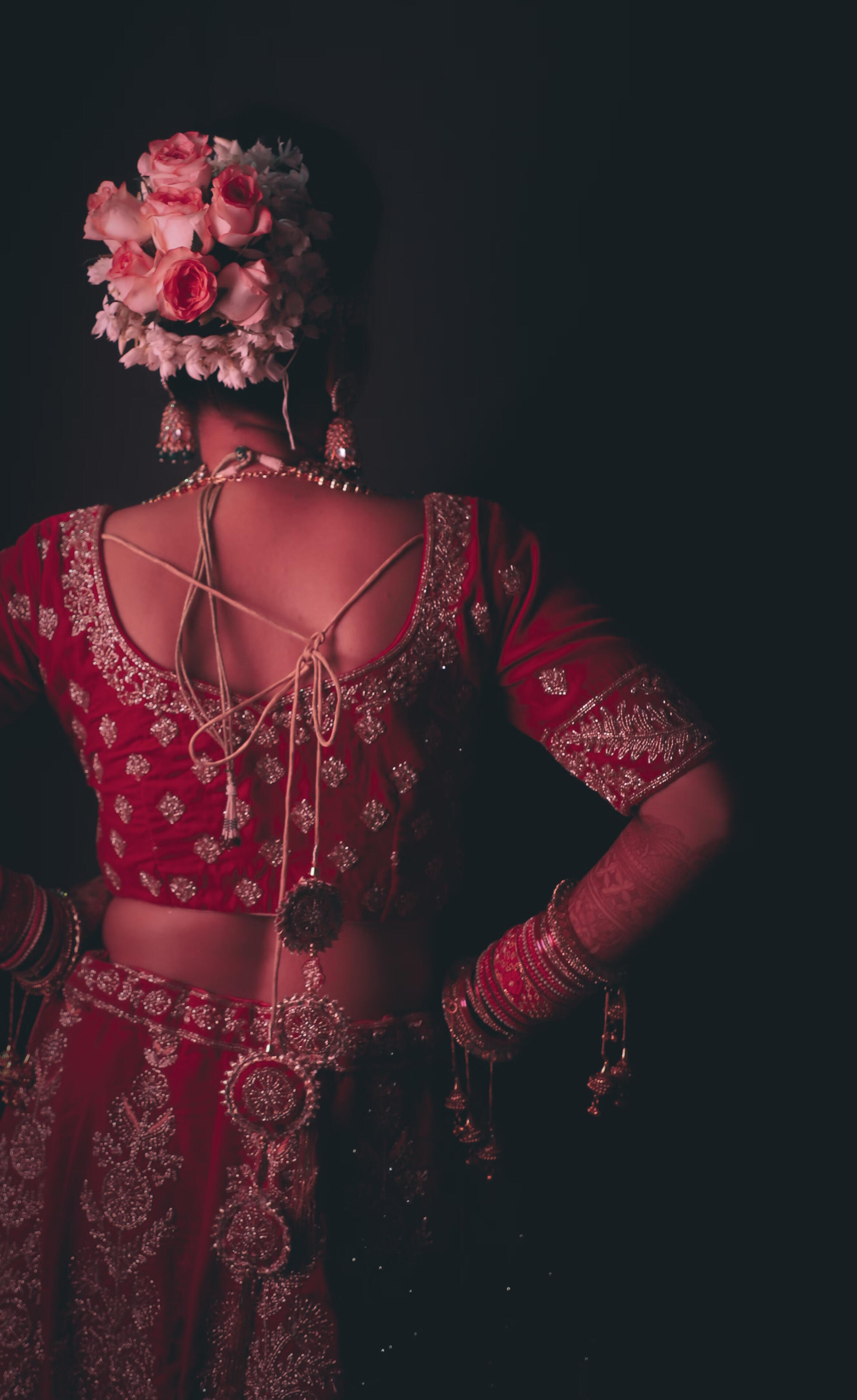 A desi woman seen from behind in a red, bridal lehenga choli with her hair up and decorated by flowers, against a black background.