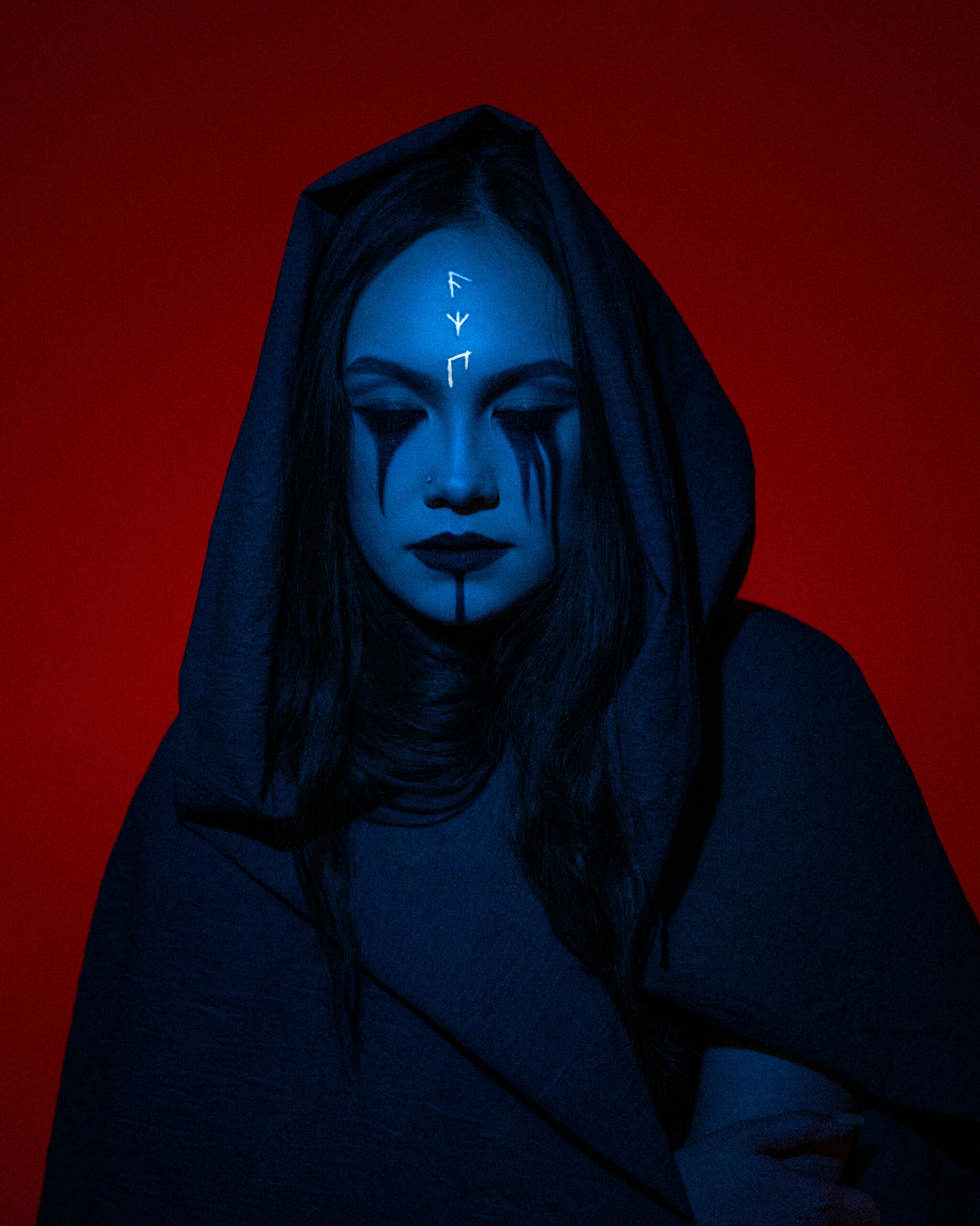 A hooded woman, eyes downcast, in bluish lighting against a red background.
